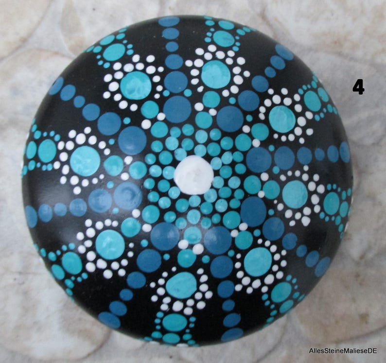 May include: A round black stone painted with a mandala pattern consisting of numerous dots in shades of turquoise, light blue, dark blue, and white. The design radiates from a single white center dot. The number 4 is visible on the right side.