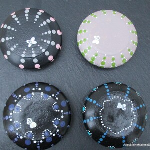 May include: Four round stones decorated with hand-painted dot patterns. One black stone features pink and grey dots, one light purple stone has green dots, one black stone has blue and white dots, and one black stone has turquoise and white dots.