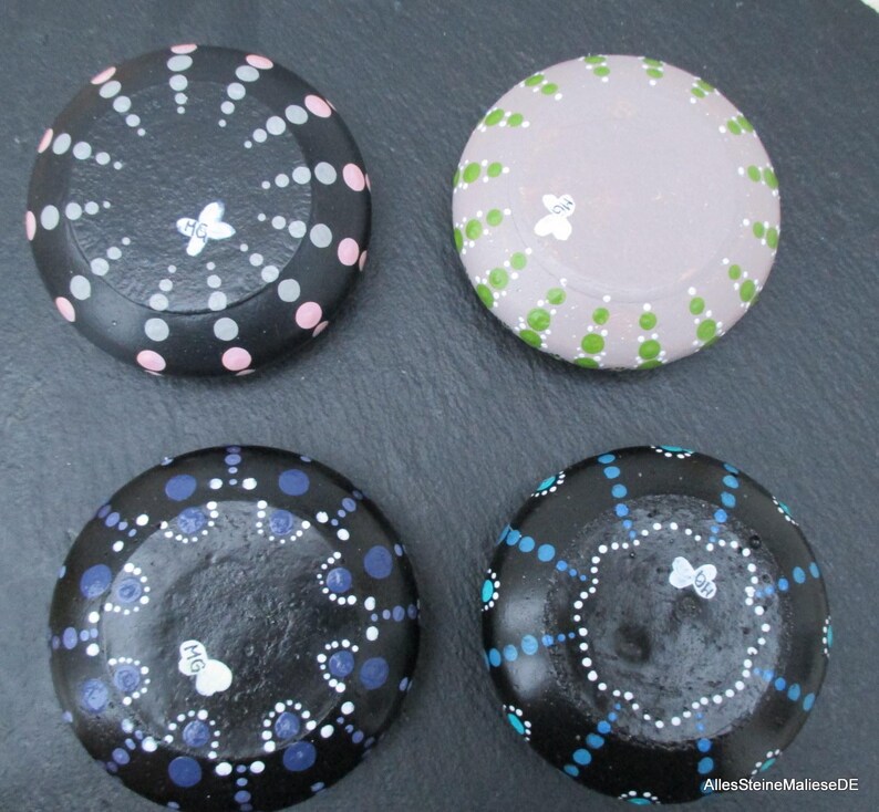 May include: Four round stones decorated with hand-painted dot patterns. One black stone features pink and grey dots, one light purple stone has green dots, one black stone has blue and white dots, and one black stone has turquoise and white dots.
