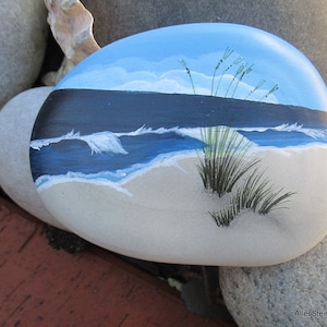 May include: A painted rock with a seascape scene. The rock is painted with a blue sky, white clouds, a dark blue ocean, white waves, a sandy beach, and green grass.