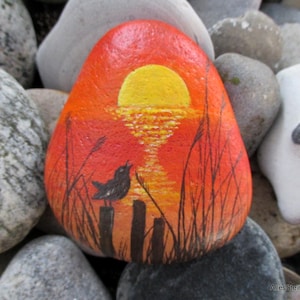 Op de afbeelding: Een gladde, afgeronde steen beschilderd met een levendige zonsondergangscène met een heldere gele zon die reflecteert op een oranje en rode horizon. Een zwart silhouet van een kleine vogel op een houten paal omringd door dunne zwarte grassprieten.