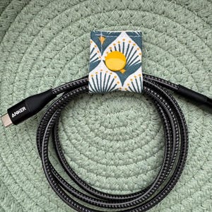 Könnte beinhalten: Ein schwarz-graues geflochtenes USB-C-Kabel mit "ANKER"-Logo, das von einem Kabelorganizer aus Stoff zusammengehalten wird. Der Organizer hat ein teal, weißes und gelbes Blumenmuster und einen gelben Knopfverschluss. Das Kabel ist aufgerollt.