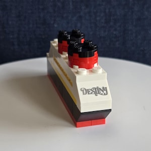 May include: A toy ship made from building blocks. The ship has a white hull with black and red details, two smokestacks, and the word "Destiny" printed on the side. It's a miniature replica of a cruise ship, constructed from colourful blocks.
