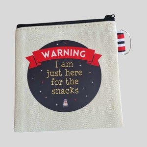 May include: A canvas zippered pouch with a black zip and a small red, white, and black striped loop. The pouch features a circular design with the text "WARNING I am just here for the snacks" in red and gold.