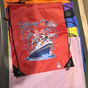 May include: A collection of drawstring bags in shades of red, orange, green, purple, blue, and pink. The red bag displays a Disney Cruise graphic with Mickey and Minnie Mouse, the text "Disney Cruise 2025", and a ship illustration.