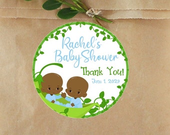 May include: A brown paper bag with a round sticker that reads "Rachel's Baby Shower Thank You!" in blue and green. The sticker features two cartoon babies in a pea pod. Greenery and a sprig of leaves are also visible.