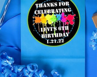 May include: A blue gift bag with a circular sticker that reads "THANKS FOR CELEBRATING LEVI'S 6TH BIRTHDAY 1.21.22". The sticker has colorful paint splatters. Blue ribbon and wrapping paper are also visible.