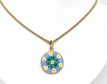 Pode incluir: Colar dourado com um pendente redondo. O pendente apresenta um design floral colorido em tons de azul, verde, amarelo e branco. A corrente é do estilo corda torcida, e o pendente é exibido em um expositor branco.