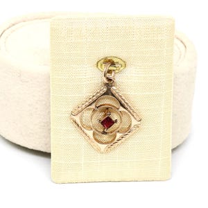 Könnte beinhalten: Ein goldfarbener Anhänger mit einem rautenförmigen Rahmen, einem komplexen durchbrochenen Design und gravierten Kanten. In der Mitte ist ein kleiner quadratisch geschliffener roter Edelstein in einem kreisförmigen Motiv gefasst. Der Anhänger hat eine goldfarbene Öse.