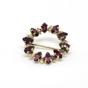 Könnte beinhalten: Eine goldfarbene Brosche mit einem runden Design, das einen Ring aus rotbraunen Steinen aufweist.
