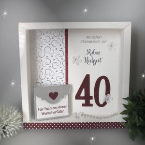 Könnte beinhalten: Ein weißer Schattenrahmen mit einem Design für einen 40. Hochzeitstag. Das Design beinhaltet die Zahl 40 in Burgunderrot, deutschen Text und einen kleinen Umschlag mit einem Herz.