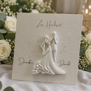 Könnte beinhalten: Eine quadratische, cremefarbene Hochzeitskarte mit einem erhabenen, weißen 3D-Relief eines tanzenden Paares. Der Text "Zur Hochzeit" ist in Braun darüber gedruckt, mit den Namen "Jennifer" und "David" in brauner Schrift an den Seiten.