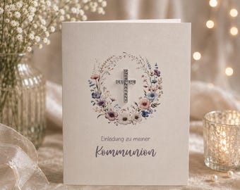 Puede incluir: Una tarjeta de invitación de color crema con una ilustración central de una corona de flores que rodea una cruz plateada. El texto dice "Einladung zu meiner" seguido de "Kommunion" en letra cursiva. La tarjeta tiene un acabado mate liso.