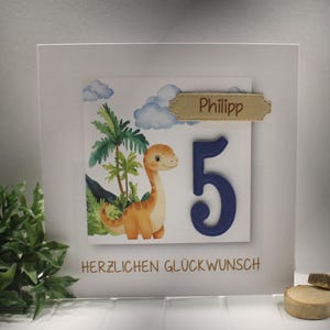 Könnte beinhalten: Eine weiße Karte mit einem Cartoon-Dinosaurier, Palmen und blauen Wolken. Die Zahl 5 ist blau, und der Name "Philipp" steht auf einem kleinen Etikett. Der Text "HERZLICHEN GLÜCKWUNSCH" ist unten aufgedruckt.