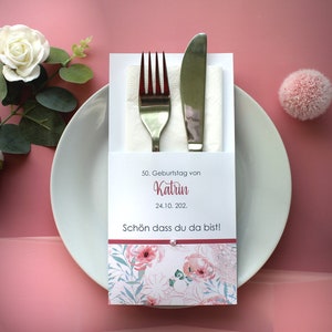 Könnte beinhalten: Eine weiße Karte mit einem floralen Design und einem roten Rand. Die Karte sagt "50. Geburtstag von Katrin 24.10.202." und "Schön dass du da bist!" Eine Gabel und ein Messer liegen in einer weißen Serviette und werden von der Karte gehalten.