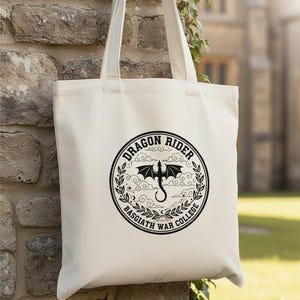 May include: Cream-coloured tote bag with a black circular graphic. The graphic features a dragon silhouette, the words "DRAGON RIDER" and "BASGIATH WAR COLLEGE" in a circular design. The bag has two long handles.