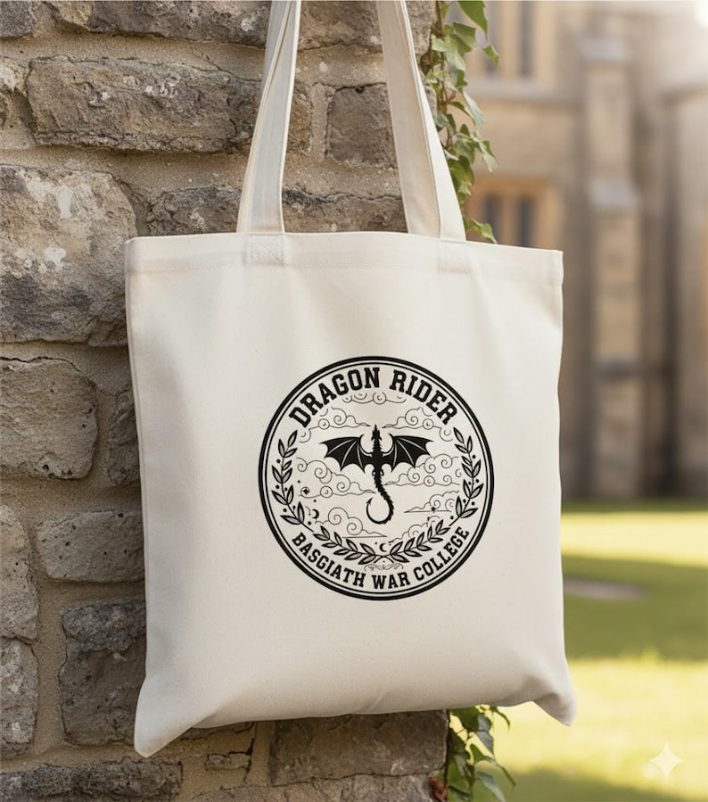 May include: Cream-coloured tote bag with a black circular graphic. The graphic features a dragon silhouette, the words "DRAGON RIDER" and "BASGIATH WAR COLLEGE" in a circular design. The bag has two long handles.