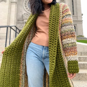 May include: A long, green and multi-colored striped crochet cardigan with a wide collar. The cardigan is open in the front and is being worn over a brown turtleneck sweater and blue jeans.