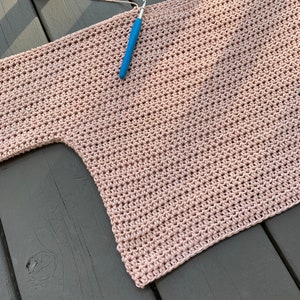 Op de afbeelding: Een roze gehaakt topje dat op een houten oppervlak ligt. Een haaknaald ligt erop en een bol garen ligt in de linkerbovenhoek.