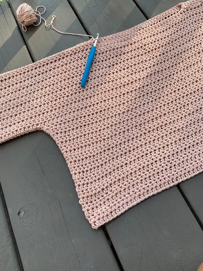Op de afbeelding: Een roze gehaakt topje dat op een houten oppervlak ligt. Een haaknaald ligt erop en een bol garen ligt in de linkerbovenhoek.
