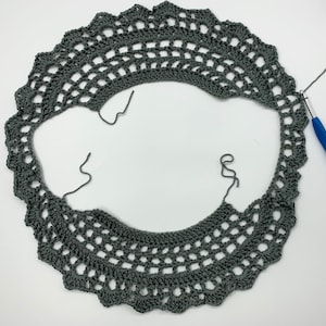 May include: A grey crocheted doily with a scalloped edge. The doily is partially finished and the crochet hook is visible.
