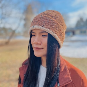 May include: A brown knit beanie with a speckled pattern and a light beige brim. The hat is made of a soft, textured yarn. The beanie is a warm accessory for cold weather.