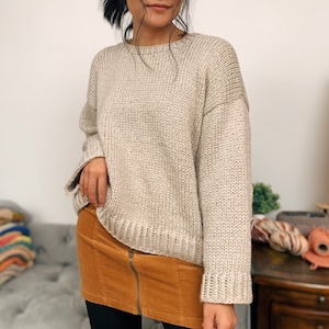 May include: A beige knitted sweater with a relaxed fit and long sleeves. The sweater is made of a soft, chunky yarn and has a slightly oversized look.