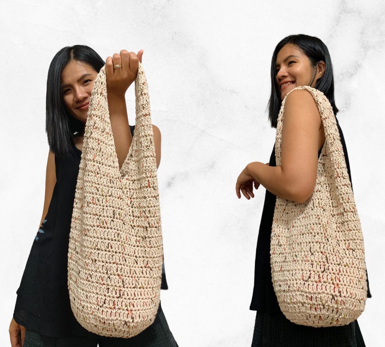 May include: A large, off-white crocheted tote bag with a speckled pattern of brown and red. The bag is worn over the shoulder of a person wearing a black tank top.