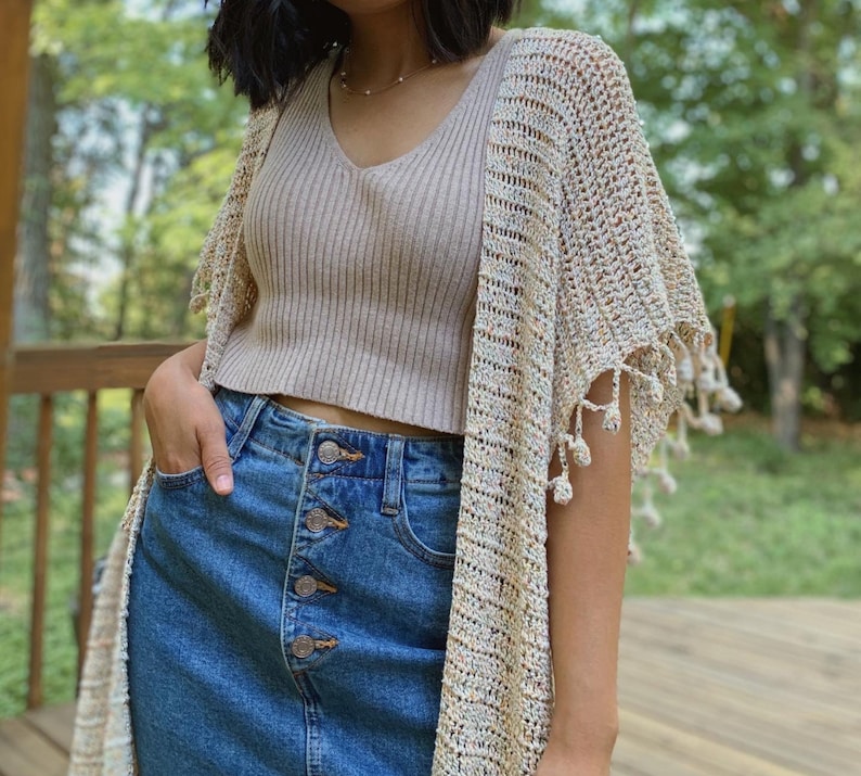 May include: A woman wearing a beige knit tank top with a crochet cardigan in a cream, brown, and white color scheme. The cardigan has fringe detailing. She is also wearing a denim skirt with button closures.