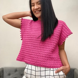 May include: A pink crocheted top with a scalloped hem and pom-pom trim. The top has a loose fit and short sleeves.