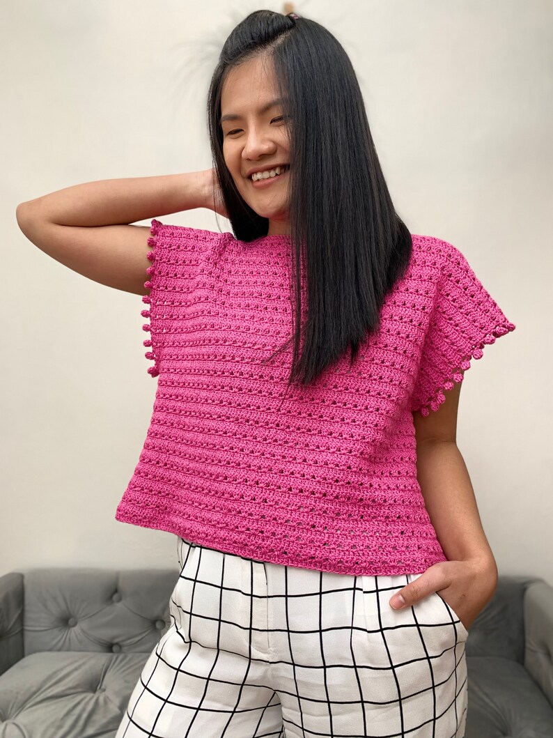 May include: A pink crocheted top with a scalloped hem and pom-pom trim. The top has a loose fit and short sleeves.