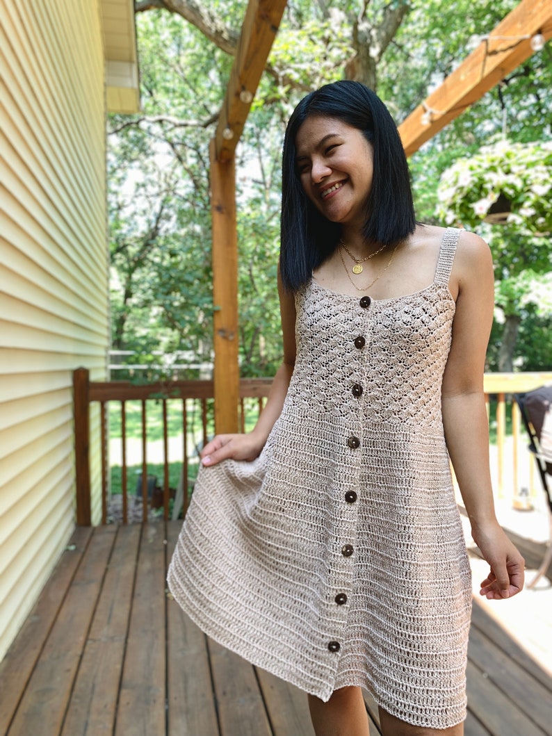 May include: A woman wearing a beige crochet dress with brown buttons. The dress has a sleeveless design and a slightly flared skirt. She is standing on a wooden deck with greenery in the background.