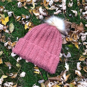 Puede incluir: Un gorro de punto rosa con un pompón blanco esponjoso. El gorro está sobre una cama de hierba verde y hojas marrones.