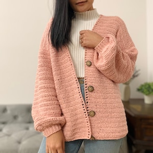 May include: A pink crocheted cardigan with a v-neck and long sleeves. The cardigan has wooden buttons and a ribbed texture.