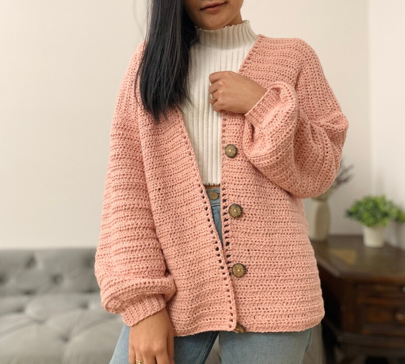 May include: A pink crocheted cardigan with a v-neck and long sleeves. The cardigan has wooden buttons and a ribbed texture.