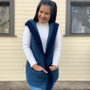 Pu&ograve; includere: Un gilet di uncinetto blu e blu navy con cappuccio e tasche. Il gilet &egrave; indossato sopra un maglione a collo alto bianco.
