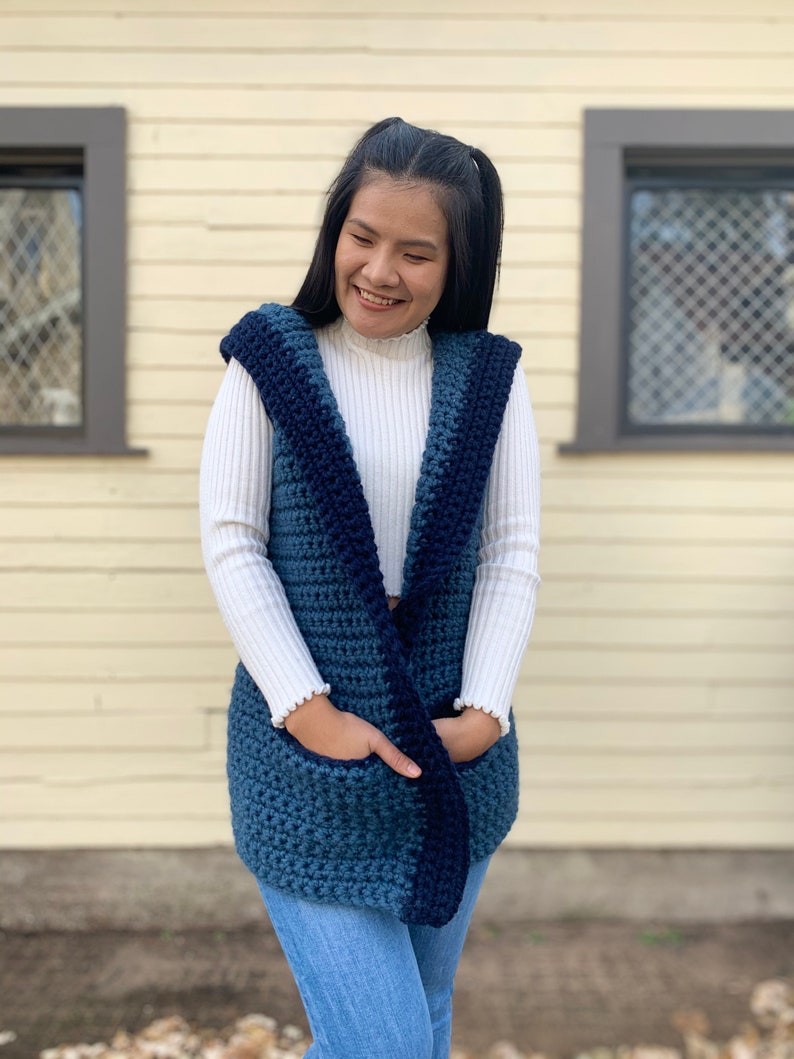 Pu&ograve; includere: Un gilet di uncinetto blu e blu navy con cappuccio e tasche. Il gilet &egrave; indossato sopra un maglione a collo alto bianco.
