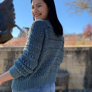 May include: A teal blue crocheted sweater with a textured stitch pattern. The sweater has long sleeves and a relaxed fit.