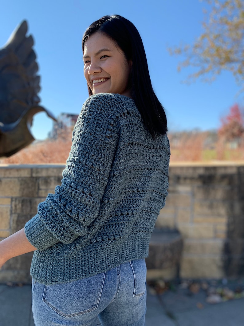 May include: A teal blue crocheted sweater with a textured stitch pattern. The sweater has long sleeves and a relaxed fit.