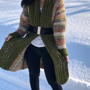 May include: A long, green crochet cardigan with a striped pattern in shades of brown, orange, and white. The cardigan has a wide collar and is belted at the waist.