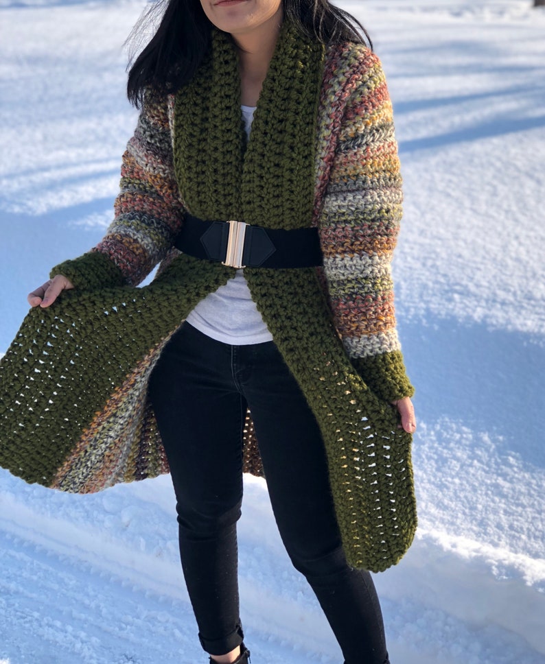 May include: A long, green crochet cardigan with a striped pattern in shades of brown, orange, and white. The cardigan has a wide collar and is belted at the waist.