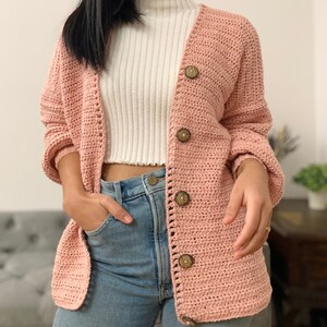 May include: A pink crocheted cardigan with wooden buttons. The cardigan has a relaxed fit and long sleeves.