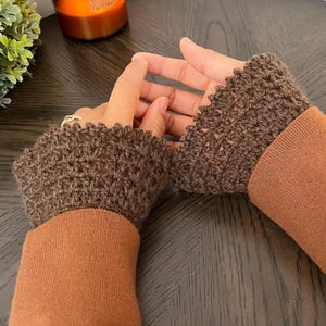 Puede incluir: Un par de guantes sin dedos de ganchillo marrones. Los guantes están hechos de un hilo texturizado y tienen un borde festoneado. Las mangas de un suéter marrón son visibles debajo de los guantes.