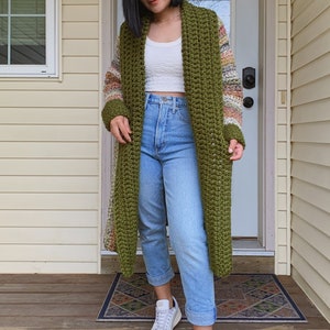 May include: A long, green crochet cardigan with a colorful striped pattern on the sleeves. The cardigan is worn over a white tank top and blue jeans. The person is wearing white sneakers.