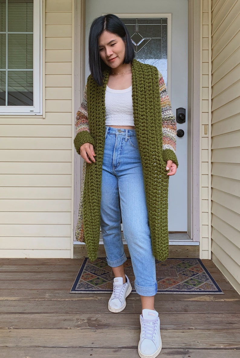 May include: A long, green crochet cardigan with a colorful striped pattern on the sleeves. The cardigan is worn over a white tank top and blue jeans. The person is wearing white sneakers.