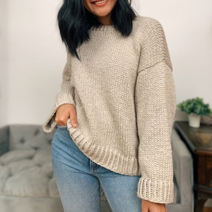 May include: A beige knitted sweater with long sleeves and a relaxed fit. The sweater has a slightly oversized look and a ribbed hem and cuffs.