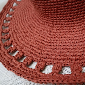 May include: Close-up of a terracotta-colored sun hat. The hat features a wide brim with a decorative, openwork edge. The woven material has a textured appearance, and the hat's crown is visible in the background.