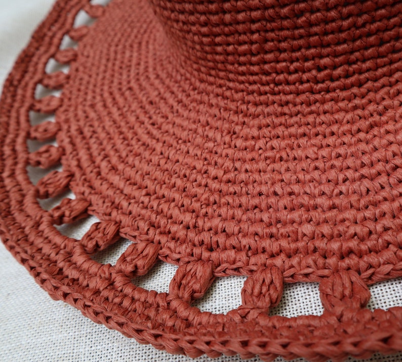 May include: Close-up of a terracotta-colored sun hat. The hat features a wide brim with a decorative, openwork edge. The woven material has a textured appearance, and the hat's crown is visible in the background.