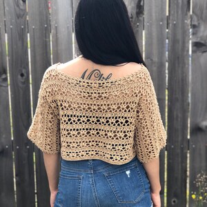 Puede incluir: Top de ganchillo color beige con mangas cortas. El dise&ntilde;o de tejido abierto del top es visible. El top se combina con pantalones cortos vaqueros azules. El fondo es una valla de madera.