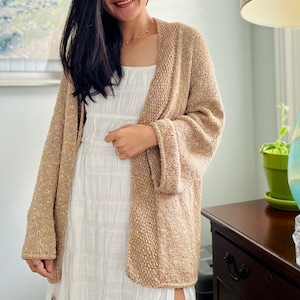 Op de afbeelding: Een beige gebreid vest met een los, open ontwerp. Het vest heeft lange mouwen en een getextureerde uitstraling met kleurvlekken. Het kledingstuk wordt over een witte jurk gedragen, waardoor een gelaagde look ontstaat. Het vest lijkt gemaakt van een zacht, comfortabel materiaal.